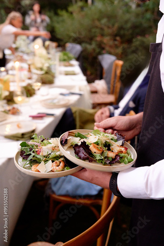 Warmly lit outdoor dinner with a long table, candles, and greens. A server carries fresh salad plates while guests mingle in the background, creating a lively, communal meal setting.