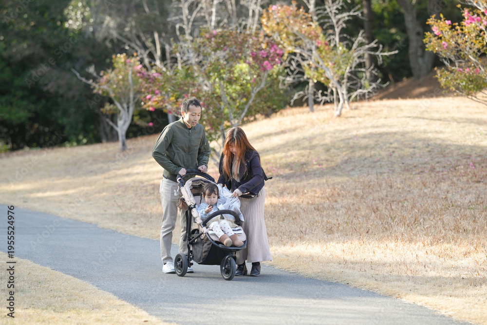Fototapeta premium Family strolls with child in a scenic park. Parents smile warmly beside their stroller. Sunlit path curves through golden grass. Trees bloom with soft pink blossoms. Perfect for parenting, family