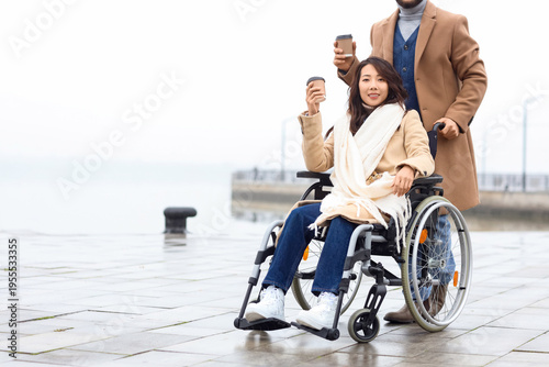 Young Asian woman in wheelchair and her husband with coffee on embankment