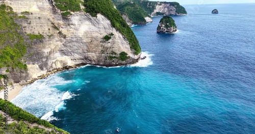 Aerial view of limestone cliffs and turquoise ocean at Kelingking Beach, Nusa Penida, Bali, with waves along a dramatic tropical coastline and offshore rock formation.