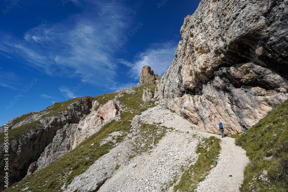 Fototapeta premium Female hiker from behind trekking alpine path with rugged cliffs under clear blue sky