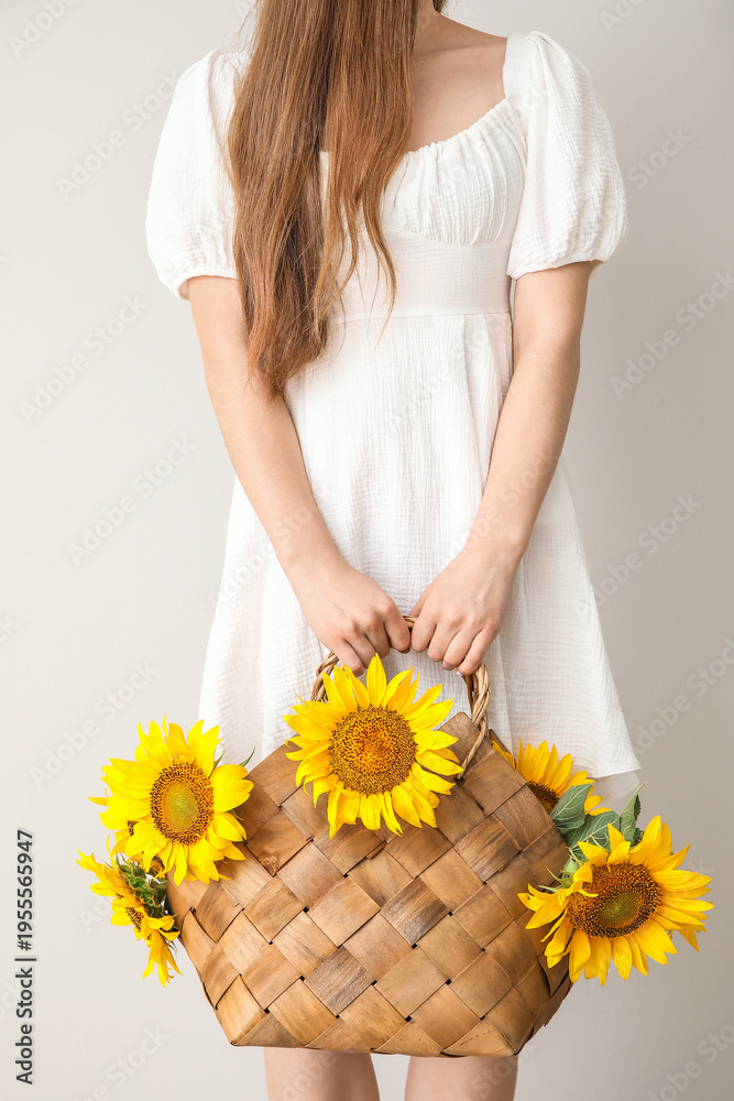 Fototapeta premium Young woman with beautiful sunflowers in wicker basket on light background