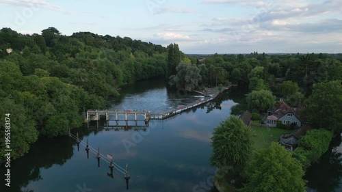 An aerial view of a tranquil river with a weir, bordered by lush green trees and a picturesque house.
