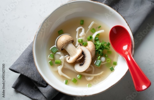 Bowl of clear oyster mushroom soup with noodles and green onions. Light asian broth with sliced mushrooms garnished with fresh herbs. Red spoon rests on side of dish.