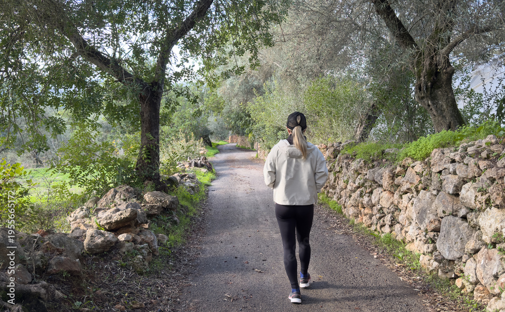 Naklejka premium A young woman seen from behind walking along a forest path, surrounded by trees and greenery. Concept of solitude, harmony with nature, outdoor lifestyle, and peaceful exploration.