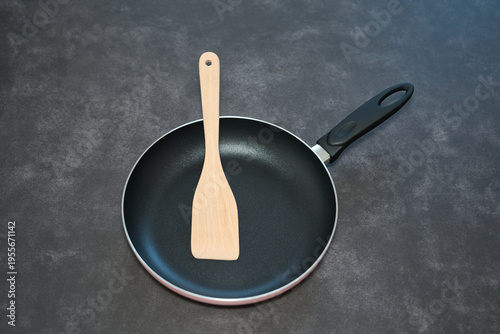 Frying pan with wooden spatula on dark surface. Nonstick frying pan with wooden spatula placed on a dark surface, showcasing essential kitchen utensils.
