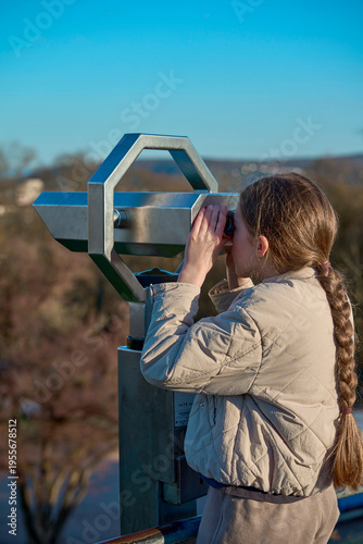 A girl with long hair on a tourist trip. A girl in a beige jacket looks through large binoculars at the beauty of the city in a tourist place.
