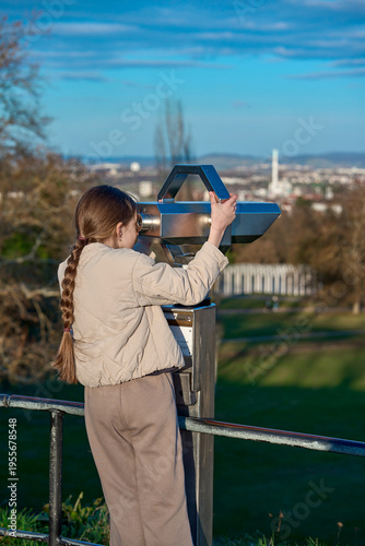 A girl in a beige jacket looks through large binoculars at the beauty of the city in a tourist place. A girl looks through binoculars on the observation deck.