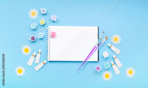 White sketchbook with paintbrushes, paints and fresh spring flowers on blue background. Creativity, artistic inspiration and celebration of World Art Day