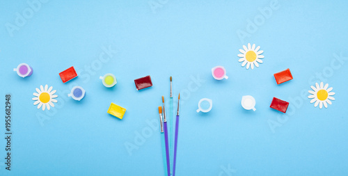 Paintbrushes with colorful paints and spring flowers on blue background. Creativity, artistic inspiration, celebration of World Art Day