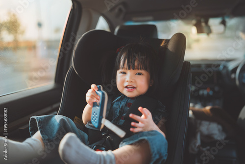 Happy little Asian girl sitting safely in car seat and looking out the window, toddler secured in child safety seat for road trip, concept of family travel, passenger safety.