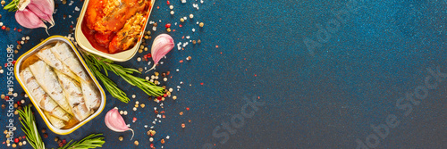 Different open tin cans with canned fish like mackerel, sprat, sardine, fish pate among spices and herbs on a blue background, viewed from the top, banner