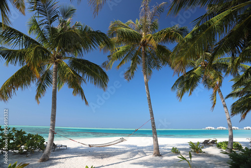 Hammock strung between coconut palm trees on white sand beach with turquoise lagoon and overwater bungalows, Maldives tropical resort