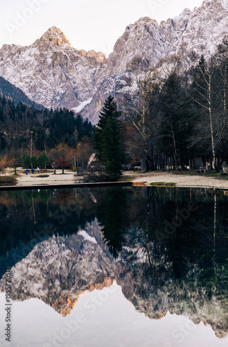 Kranjska Gora, Slovenia - November 23, 2023: Scenic view of snow-capped mountains reflecting in a tranquil lake surrounded by trees and park pathways in a natural landscape