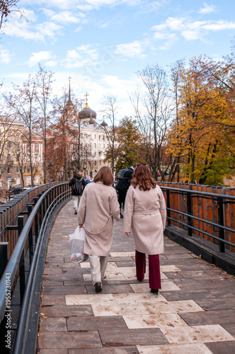 Two women walking across a bridge on an autumn day, seen from behind, wearing beige coats