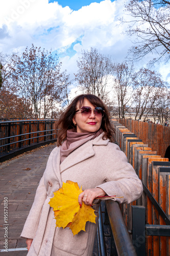 Woman holding a large yellow leaf while posing for a photograph taken by another woman in a cozy outdoor setting during autumn, showcasing seasonal beauty and friendship