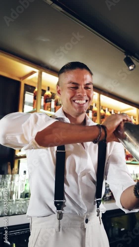 Professional bartender smiling and shaking a stainless steel shaker. Young mixologist making a drink behind the bar in a stylish establishment