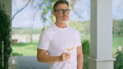 Mature man drinking glass of white wine on terrace of farmhouse home, embracing tranquility an peace of countryside. Satisfied landowner looking, overviewing ranch, enjoying rural lifestyle.