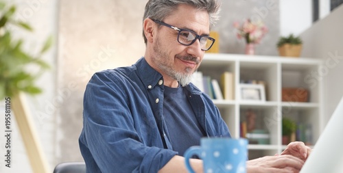 Happy man with laptop computer at home. Businessman working at desk in home office. Portrait of mature age, middle age, mid adult man, bearded, glasses, smiling, authentic look.