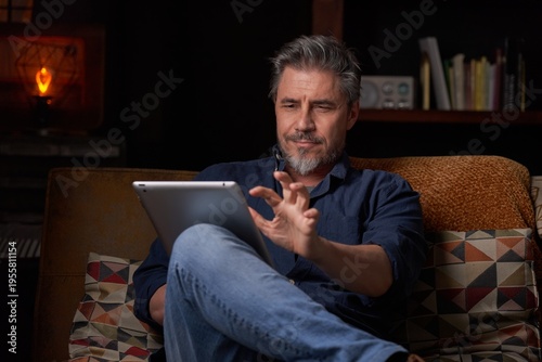 Man sitting on couch at home with tablet computer. Businessman working in home office. Portrait of mature age, middle age, mid adult man, bearded, authentic look.