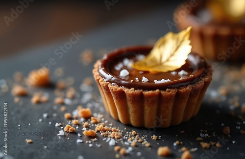 Mini chocolate tart with salted caramel filling sits on dark surface. Topped with gold leaf, sea salt flakes, and pastry crumbs scattered around the delicious sweet.