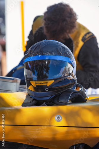 Black and Yellow Racing Helmet with Blue Tape Resting on a Vehicle while a person in a racing vest works in the blurred background.
