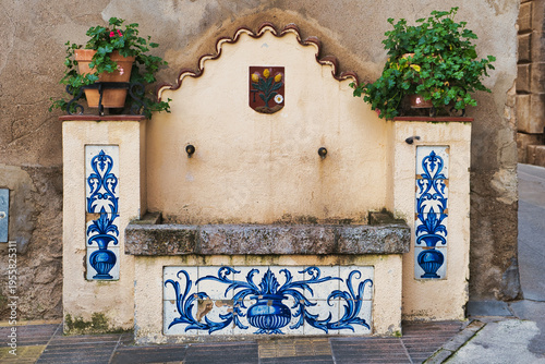 Old, well-used village fountain, decorated by blue ceramic tile panels and potted plants, in the Spanish town of Tivissa, Catalonia
