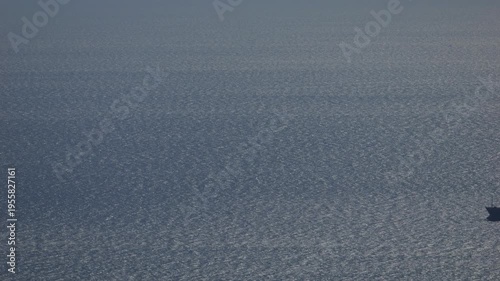 Telephoto view of a cargo ship moving across a shimmering sea surface under soft cloud shadows. Minimalist maritime scene with vast negative space and subtle light variations, ideal for themes of glob
