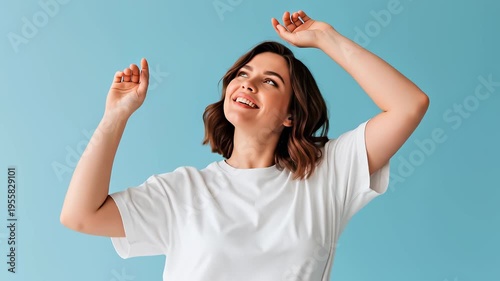 Smiling young woman in white t-shirt dancing with raised arms on blue background