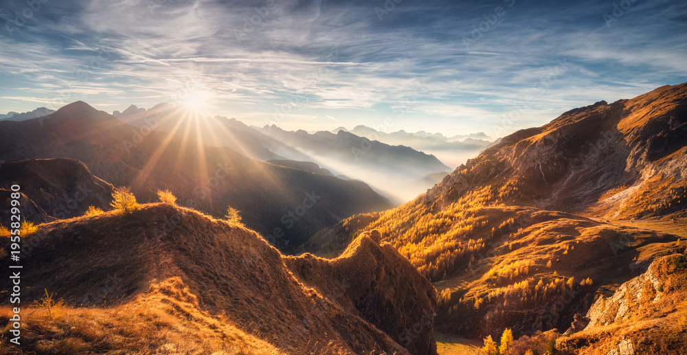Naklejka premium Mountains in low clouds at beautiful sunset in autumn in Dolomites, Italy. Landscape with alpine mountains, orange grass and trees, hills in fog, blue sky with clouds and sun in fall. Nature