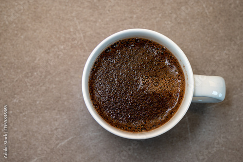 Top down view of a white ceramic mug filled with fresh black coffee showing natural crema and bubbles. Minimalist lifestyle shot on a neutral stone background with copy space.