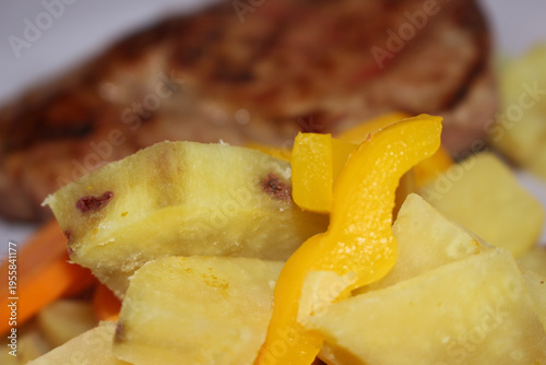 Cooked sweet potato with bell pepper and carrots. Steamed vegetables served with steak in background. Healthy boiled veggies and beef meal.