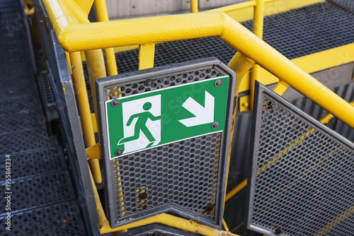 Industrial emergency exit sign with arrow direction. Safety signage mounted on metal structure in factory environment. Evacuation route indicator for workplace security and hazard protection.