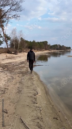 Man walking alone on a sandy beach during a cloudy spring day, reflecting in calm water. Mindful walk, thinking and rest