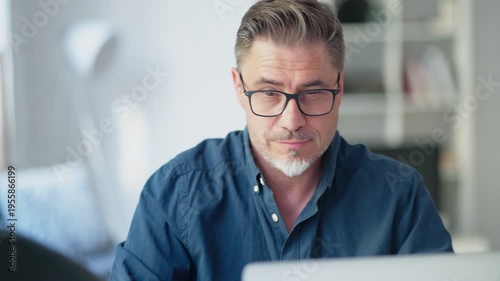 Older man sitting at desk in bright room working on laptop computer in home office. Mature age, middle age, mid adult casual man in 50s, confident happy smiling.