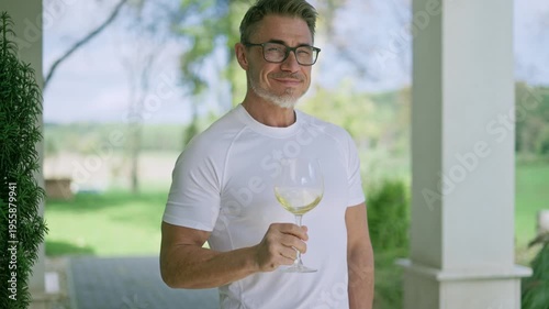 Mature man drinking white wine on porch of country house. Happy middle aged male in white outfit standing outside on ranch terrace tasting from glass, smiling. Tranquility and countryside lifestyle.