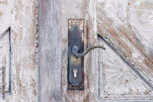 Old weathered wooden door with vintage metal handle and keyhole. Rustic entrance detail with peeling paint and antique hardware. Aged texture showing history and character of traditional architecture.