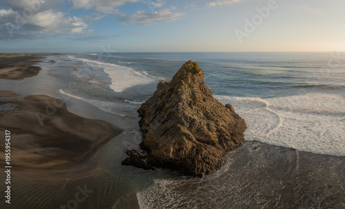 Aerial view of the iconic Rock at Karekare Beach, New Zealand. Waves crash around the rock formation, showcasing the rugged beauty of the coastline in Auckland.