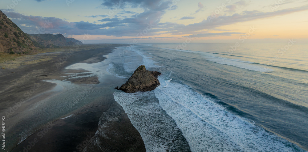 Fototapeta premium Aerial view of the black sands of Karekare Beach, New Zealand, with a Rock standing tall against the crashing waves. A popular spot for surfers and nature lovers.