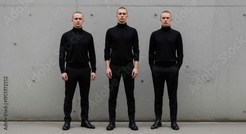 Three men in black turtlenecks and pants stand against a gray wall.