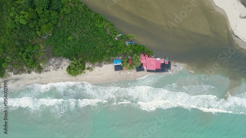 Drone view of red-roof beach huts and a blue boat as turquoise surf and brown river water mix along a narrow sandy shoreline in Palomino.