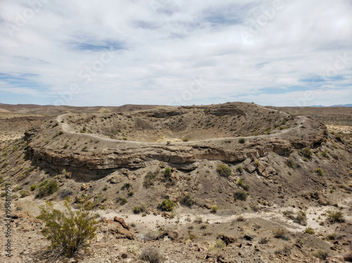 A wide, arid landscape featuring a large, circular impact crater with rugged, rocky walls under a bright, cloudy sky.