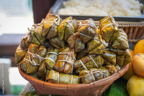 A pile of Khao Tom Mat (sweetened sticky rice with banana and black beans) wrapped in green banana leaves and tied with bamboo strips, displayed in a wicker basket at a Thai street food market.