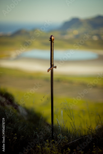 A medieval iron sword is stuck into the ground against a backdrop of mountains and the sea. Vertical photo