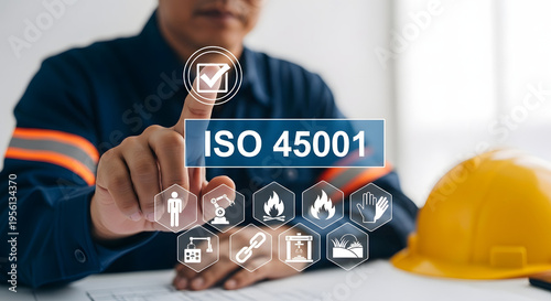 A worker in a blue uniform interacts with a virtual interface displaying ISO 45001 icons on a table next to a yellow safety helmet.