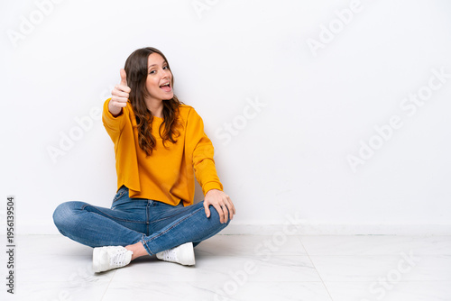Young caucasian woman sitting on the floor isolated on white wall with thumbs up because something good has happened