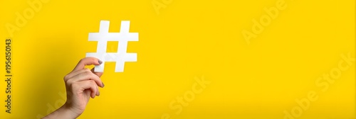 A human hand gently holds a crisp white paper hashtag symbol against a bright and cheerful yellow background under soft even studio lighting creating a clean modern and conceptual digital media repres