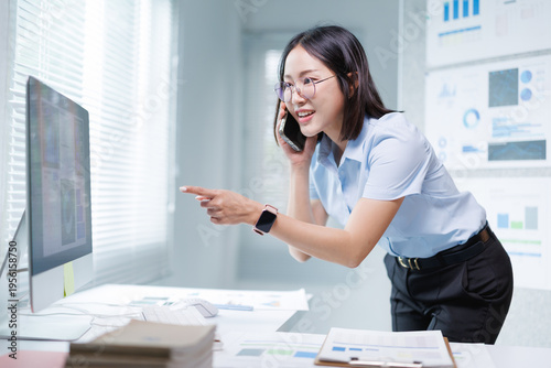 Asian businesswoman talking on a mobile phone while working in a modern office, standing and pointing at content on a desktop computer screen during a busy workday
