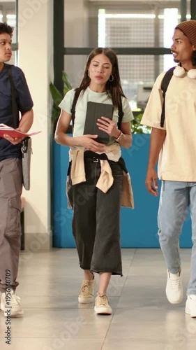 Vertical video: Walking student with tablet, in green tee, heading to class in hall with red folder