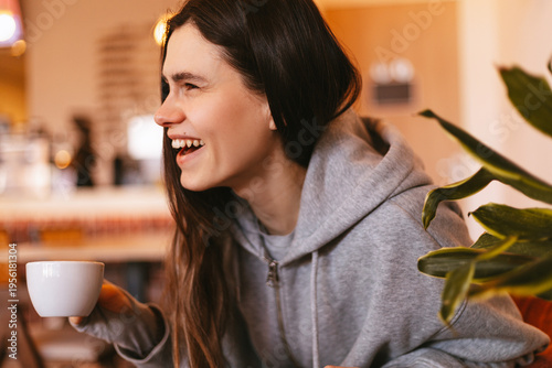 Fotografie Side view of happy laughing young woman in grey hoodie holding white cup of coff